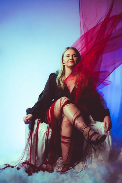 Studioportrait einer blonden Frau mit fließendem roten Stoff vor blauem Hintergrund.