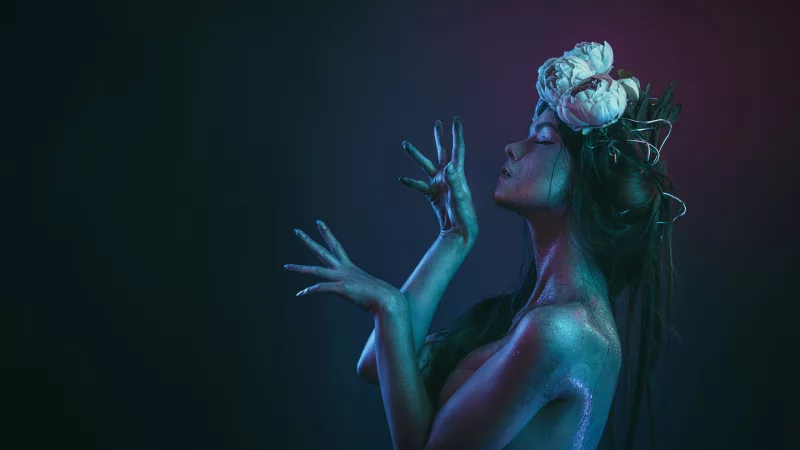 Studioportrait einer Frau mit glitzernder Haut und weißen Blüten im Haar unter farbigem Licht.