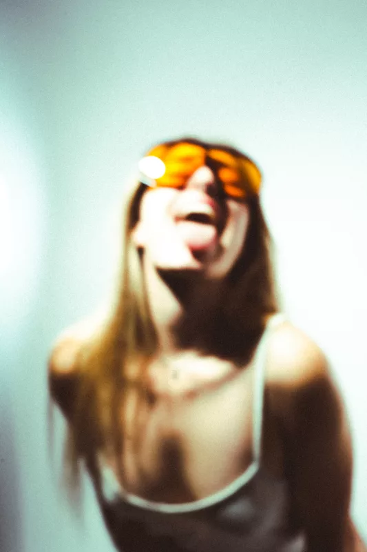 Unscharfes Porträt einer Person mit orangefarbener Sonnenbrille und herausgestreckter Zunge