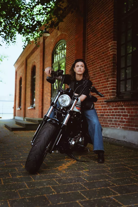 Urbanes Portrait einer Frau auf einem schwarzen Motorrad vor Backsteinarchitektur