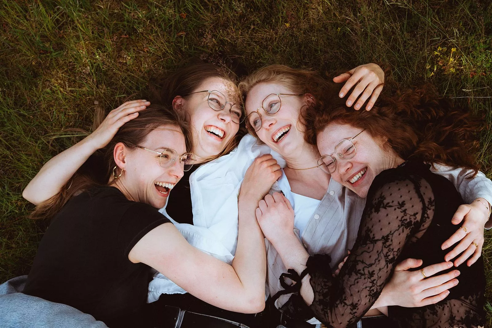 Vier lachende Frauen mit Brillen liegen gemeinsam im Gras.
