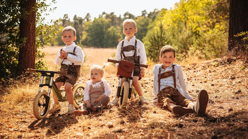 Vier kleine Jungen in rustikaler Kleidung mit Laufrädern auf einer sonnendurchfluteten Waldlichtung.