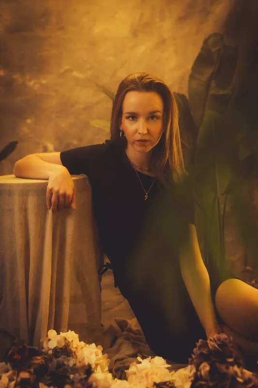 Warm beleuchtetes Studioportrait einer Frau in einem schwarzen Kleid vor tropischen Pflanzen.