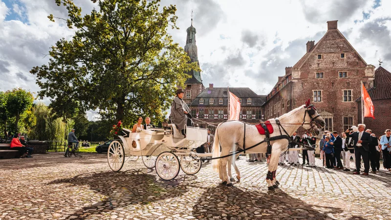 Weiße Pferdekutsche auf Kopfsteinpflaster vor historischen Backsteingebäuden und einem Kirchturm.