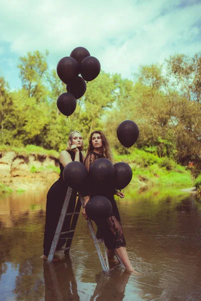 Zwei Frauen in dunklen Kleidern halten eine Leiter mit schwarzen Luftballons im flachen Wasser.