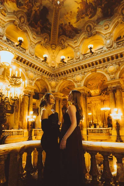 Zwei Frauen in eleganten schwarzen Kleidern in einem prachtvollen, goldenen Saal.
