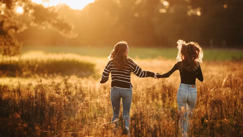 Zwei Frauen gehen Hand in Hand durch ein hohes Grasfeld im warmen Abendlicht.