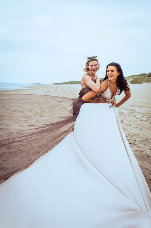Zwei lachende Frauen in fließenden Kleidern an einem Sandstrand