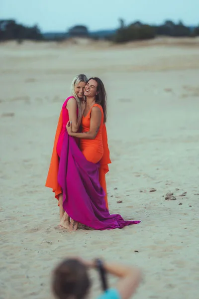 Zwei lachende Frauen in leuchtend orangefarbenen und pinken Kleidern am Strand.